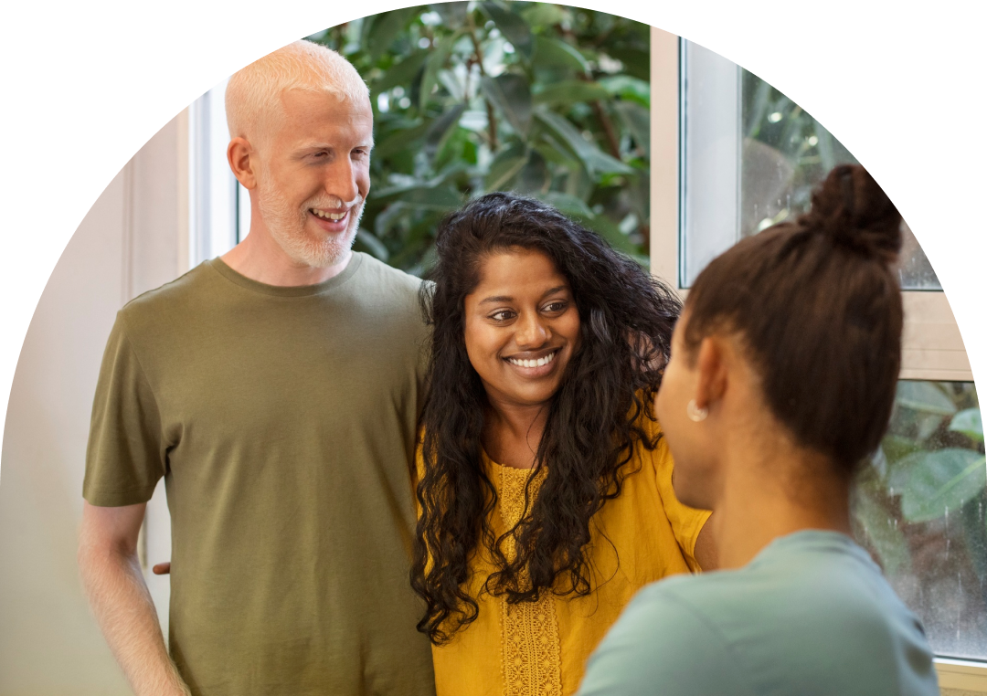 Three adults standing indoors near a window, smiling and talking together. A man with short gray hair stands beside a woman with long dark hair, both looking at another woman in the foreground. Green plants are visible outside the window.
